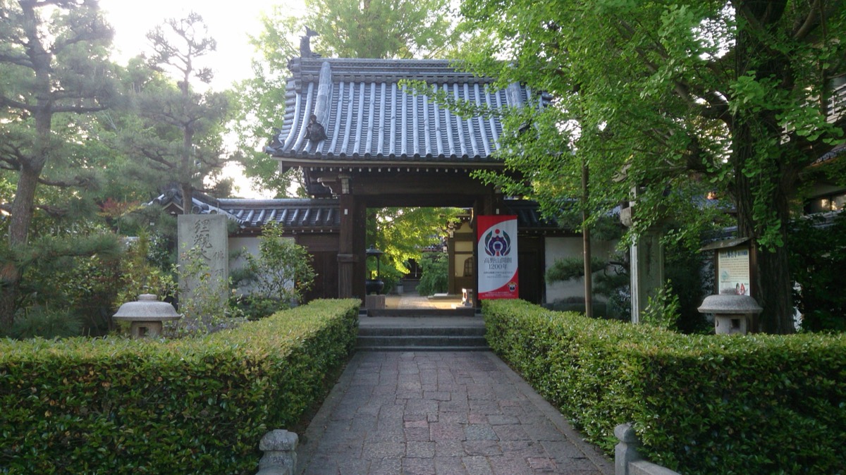 양옆으로 다듬은 산울타리가 이어진 돌길 끝에 기와지붕을 얹은 사찰 산문이 서 있고, 문 안쪽에 붉고 흰 깃발이 걸려 있다. 왼쪽에 돌기둥과 석등, 오른쪽에 큰 은행나무와 안내판이 보인다